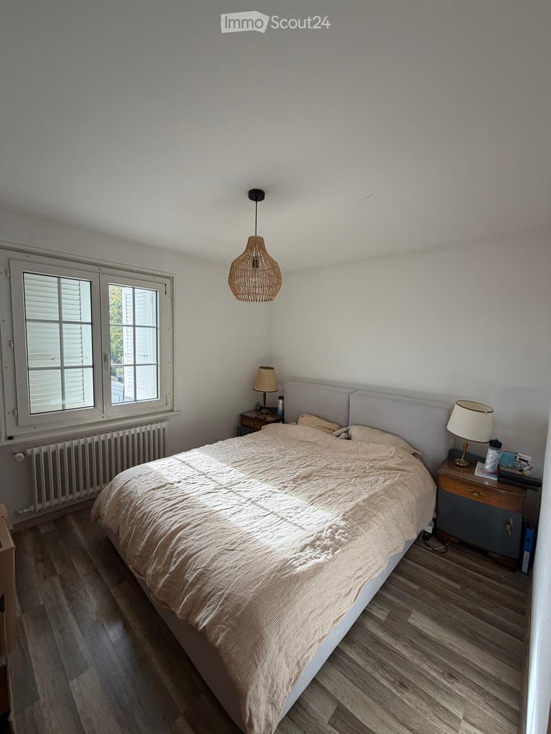 Cozy bedroom with wooden flooring, a large bed with beige bedding, two nightstands with lamps, a wicker pendant light, and a radiator beneath a window with white shutters.