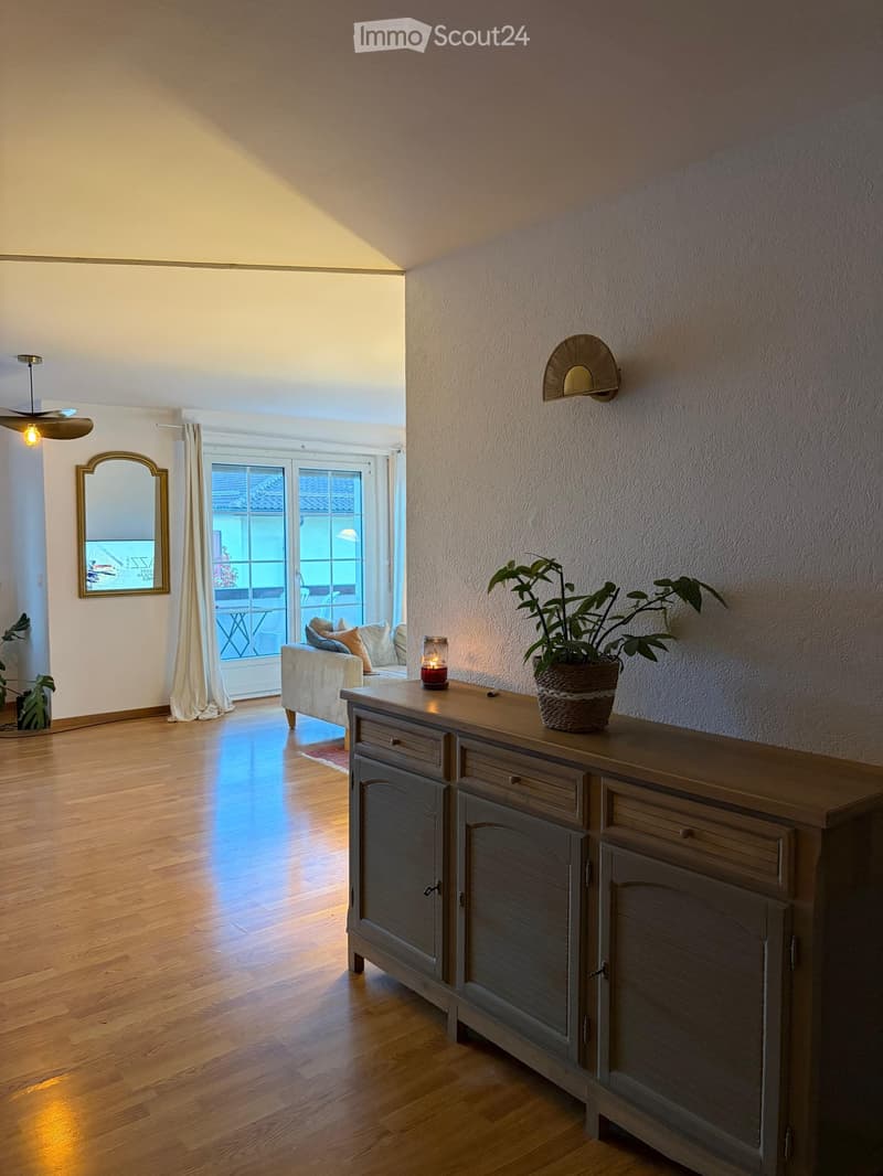 A well-lit living room with wooden flooring, a sofa with cushions, a wooden cabinet with drawers, a potted plant, a lit candle, a wall-mounted lamp, and a large mirror on the wall.