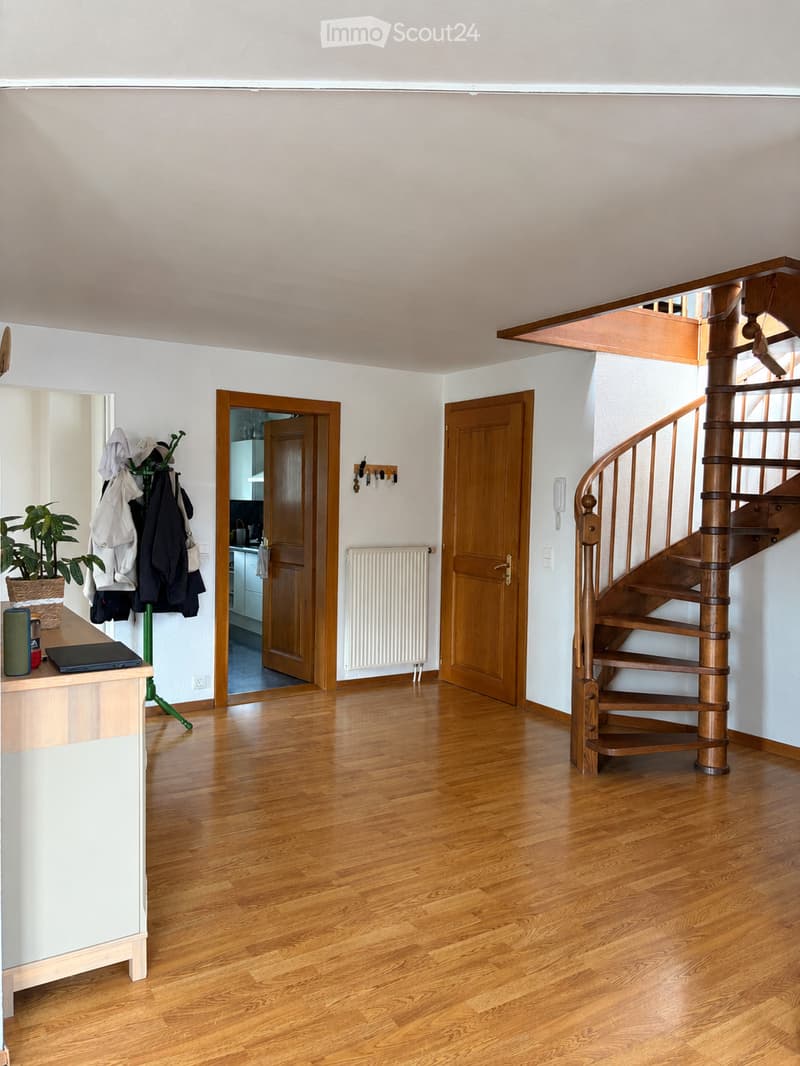 Spacious hallway with wooden flooring, a spiral staircase, and several doors leading to other rooms. A coat rack with jackets and a small plant is visible on the left side.