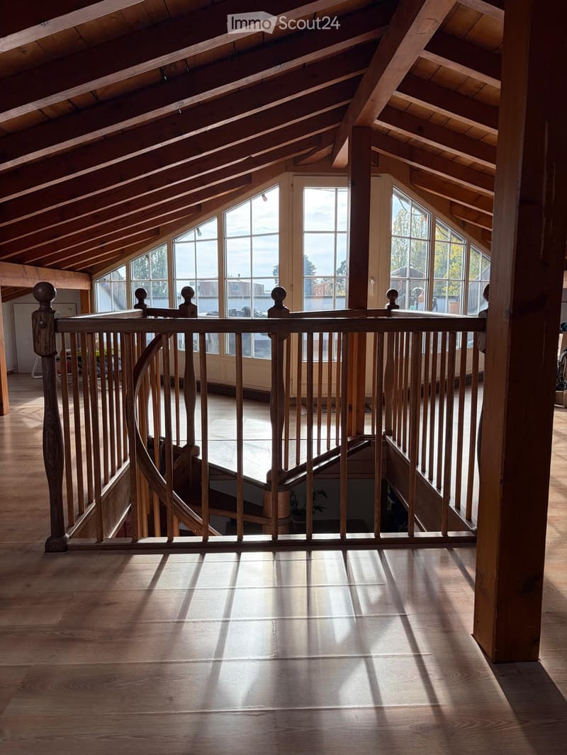 Spacious, high-ceilinged hallway with wooden beams, large windows, and a wooden railing leading to a lower level.