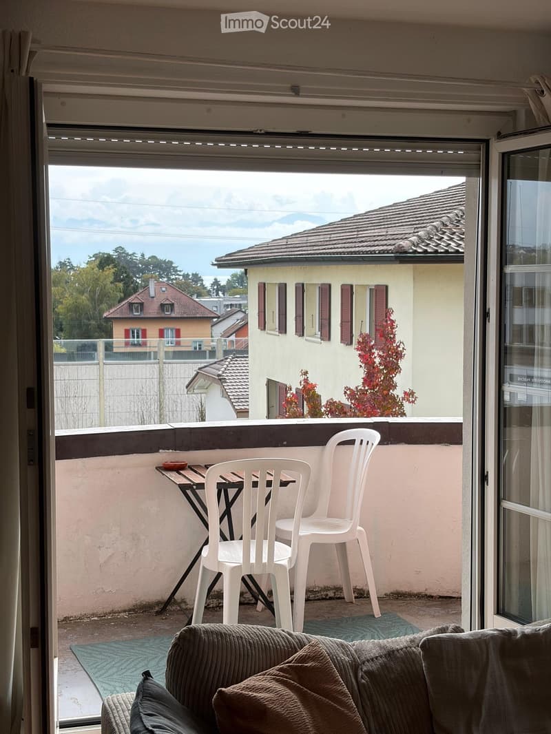 Balcony with white plastic chairs and a small table, overlooking a residential area with houses and trees.