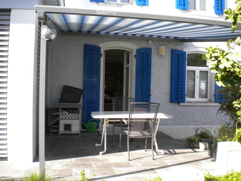 A covered patio with a white table and two metal chairs, a green watering can, and potted plants. The patio has a striped awning and is adjacent to a house with blue shutters.
