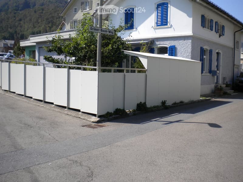 Two-story residential building with white walls, blue shutters, and a covered entrance. A white fence lines the front yard, and the street sign reads "Grüner-Baum-Weg."