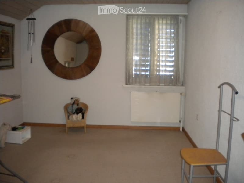 A small, furnished room with a large round mirror on the wall, a window with curtains, a small chair with a stuffed animal, a radiator, and a stool near the door.