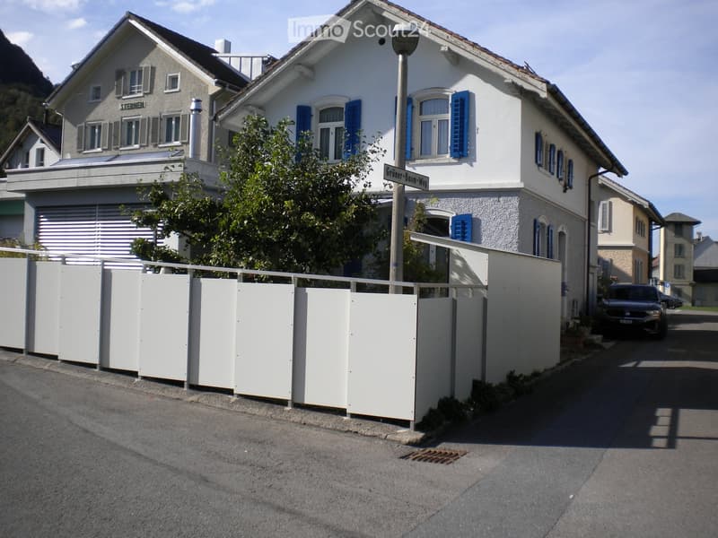 Two-story residential house with white walls, blue shutters, and a garage. A white fence surrounds the property, and a car is parked nearby.