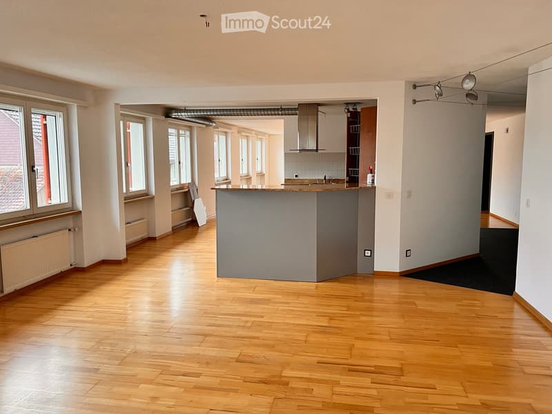 Empty room with wooden flooring, large windows, and a kitchen area visible in the background. No furniture or appliances are present.