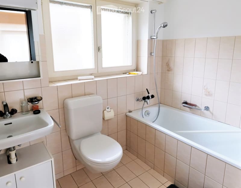 Bathroom with a toilet, bathtub, sink, and mirror. Light-colored tiles and a window provide natural light.