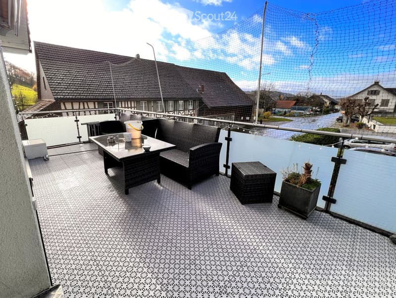 Balcony with a wicker sofa, table, stool, and potted plant, overlooking a residential street.