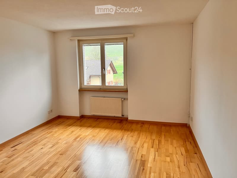 Empty room with wooden flooring, white walls, a window with a view of a house and greenery, and a radiator below the window.