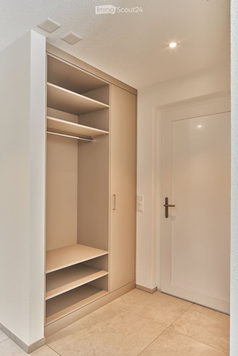 Empty room with beige tiled floor, white walls, built-in closet with multiple shelves and a hanging rod, and a closed white door.