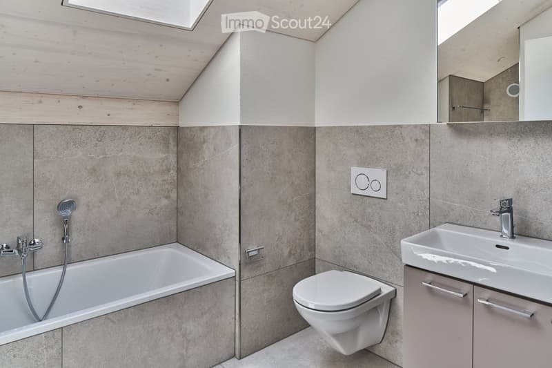 Modern bathroom with a bathtub, toilet, sink, and mirror. Features grey tiled walls and a skylight window.