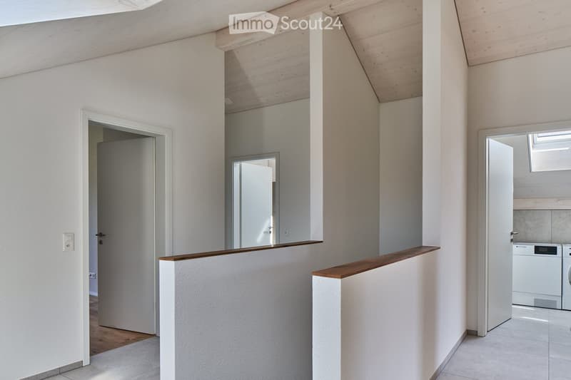 Bright, modern hallway with white walls, wooden ceiling beams, and a wooden railing leading to a laundry area with a washing machine and dryer.