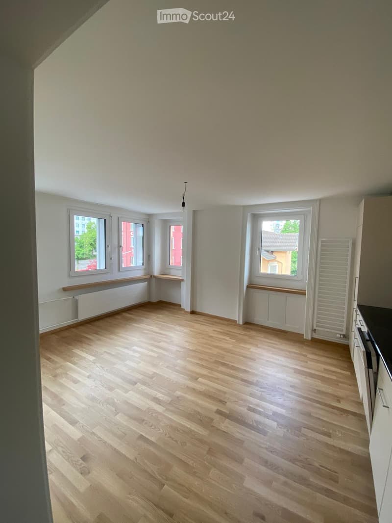 Empty room with light wooden flooring, white walls, and multiple windows. Radiators are visible beneath the windows, and there is a small section of a kitchen counter visible on the right side.