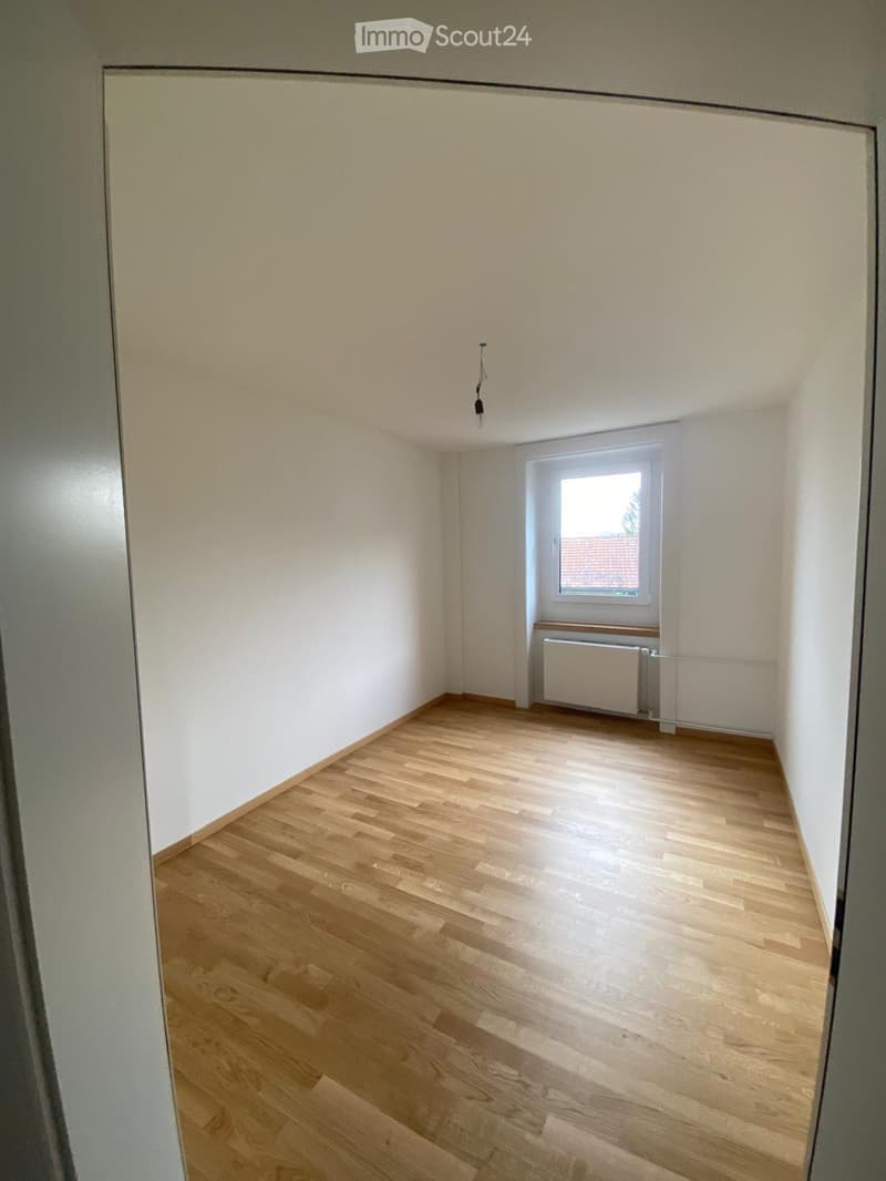Empty room with light wooden flooring, white walls, and a single window with a radiator below it.