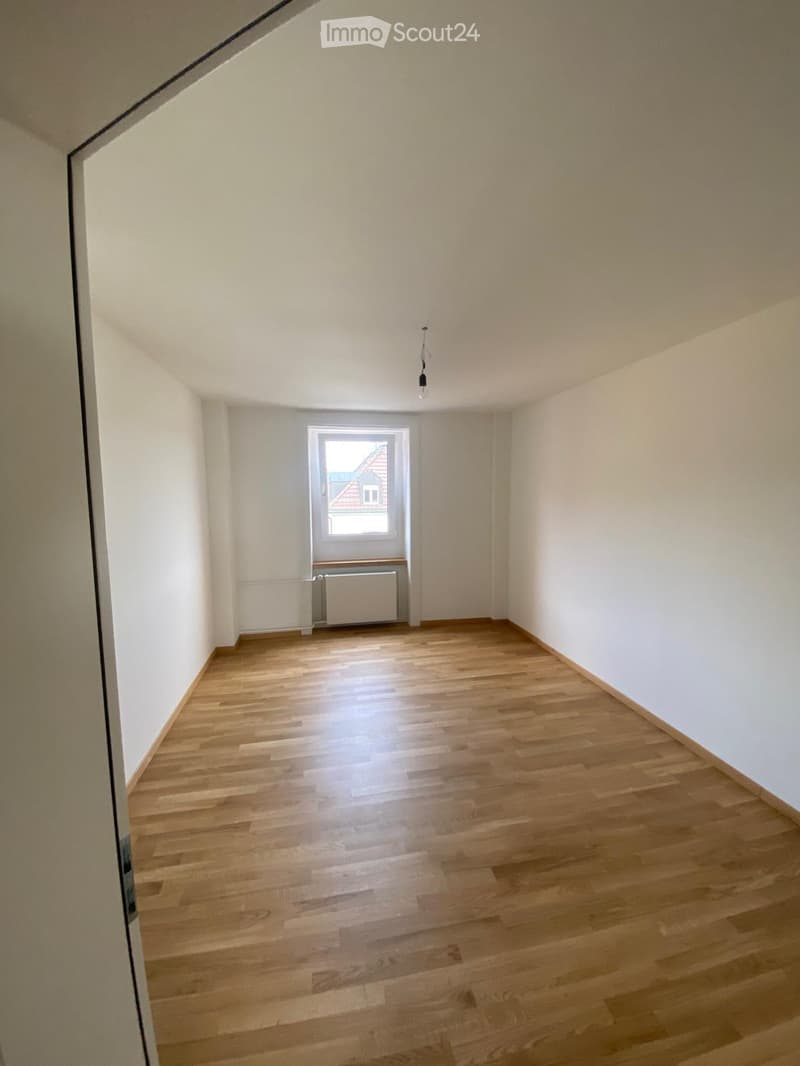 Empty room with wooden flooring, white walls, and a single window showing a view of a neighboring building.