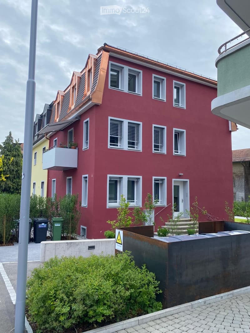 Red two-story house with white-framed windows, a balcony on the upper floor, and a small garden area in front.