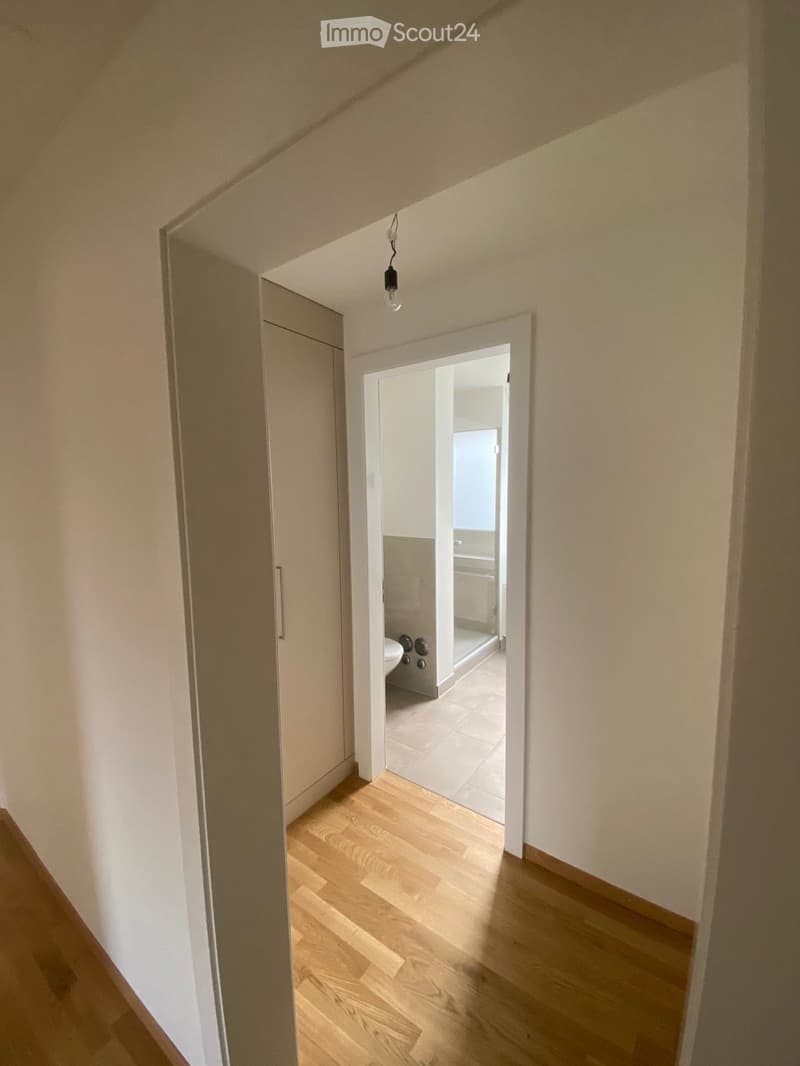 A narrow hallway with light wooden flooring and white walls, leading to an open doorway that reveals a small bathroom with a toilet and tiled floor.