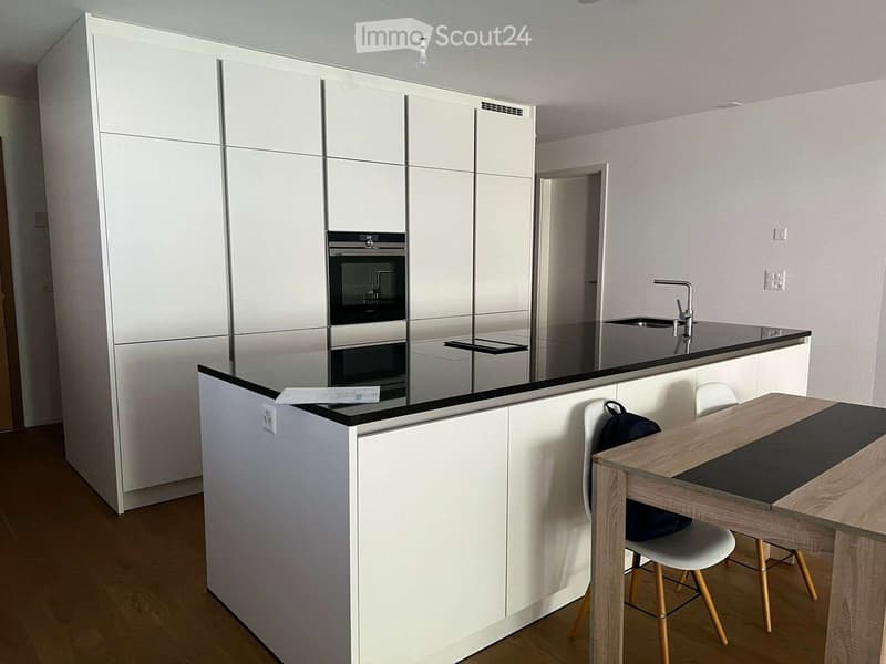 Modern kitchen with white cabinets, a central island, a wooden dining table with chairs, and built-in appliances including an oven and sink.