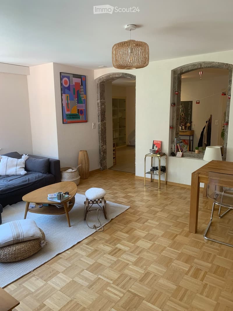 Living room with wooden herringbone flooring, a dark blue sofa with white pillows, a round wooden coffee table, a small round stool, a wicker chair, a small side table with a lamp, and a large woven pendant light fixture.