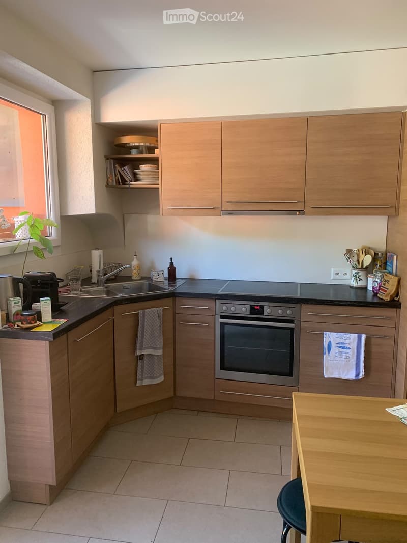 Modern kitchen with light wooden cabinets, stainless steel appliances, and a black countertop. Includes a sink, oven, and a small dining area with a table and chair.