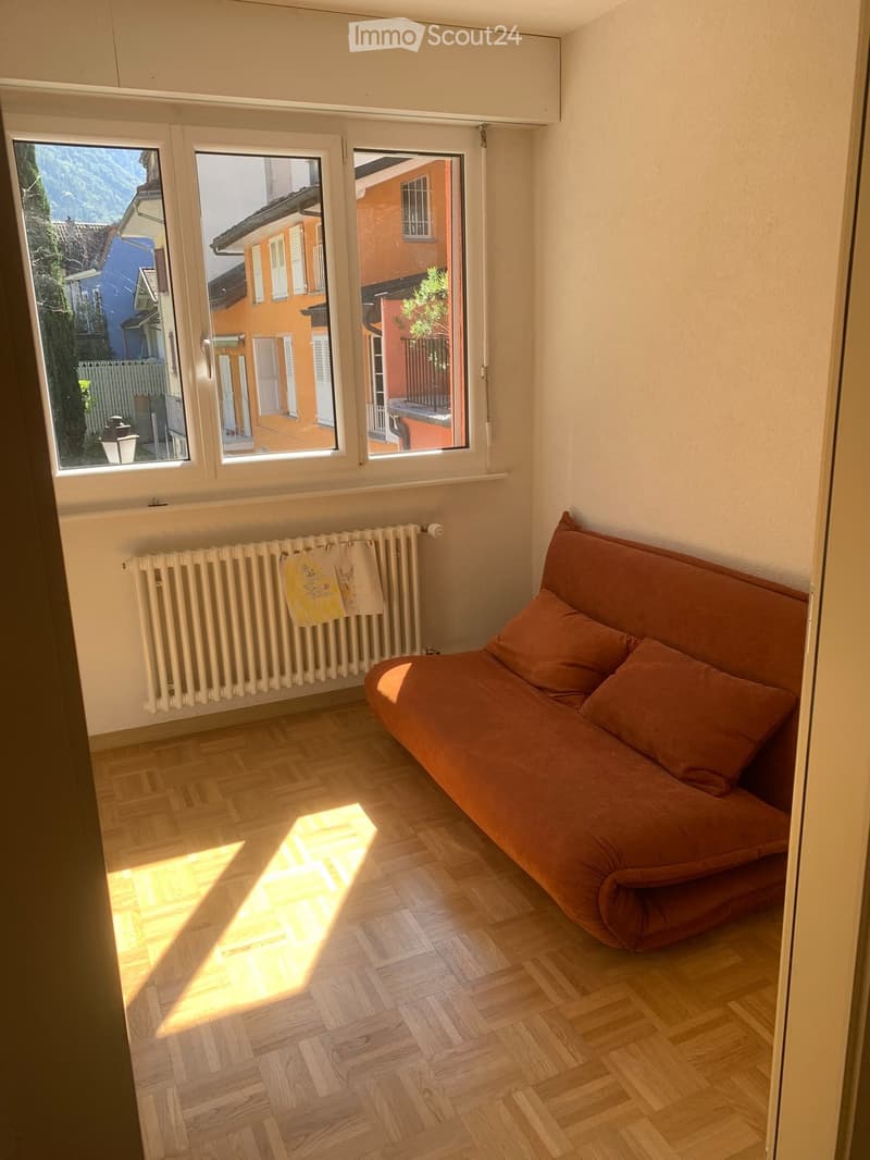 Living room with wooden flooring, a brown sofa with orange cushions, and a radiator with a towel hanging on it. Large windows offer a view of the street and surrounding buildings.