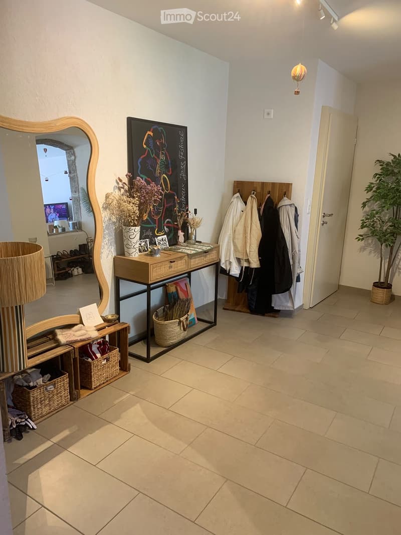 Hallway with tiled floor, coat rack, small table with decorative items, mirror, and a view into another room with a TV and plant.