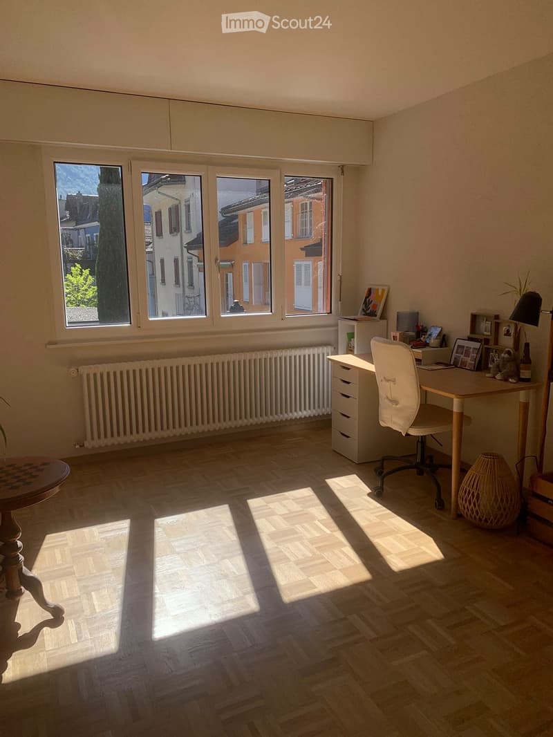 Bright room with wooden flooring, large windows, desk with chair, small table, lamp, radiator, and decorative items.