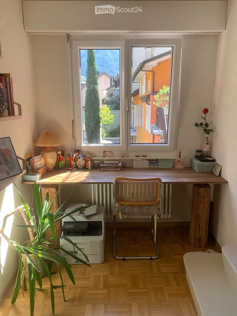 Brightly lit room with wooden flooring, large windows, a desk with a chair, a printer, a lamp, and decorative items including a plant and picture frames.