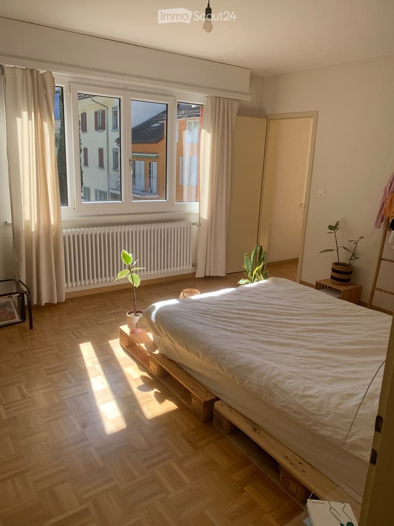 Bright bedroom with wooden herringbone flooring, large windows with white curtains, a bed on a wooden platform, a radiator, small tables, and potted plants.