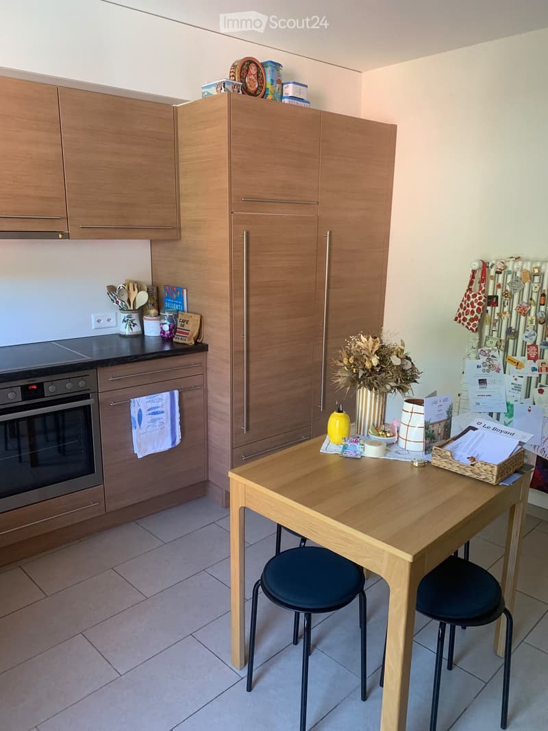Modern kitchen with wooden cabinets, stainless steel oven, and a wooden table with two stools. The counter has various kitchen utensils, books, and decorative items.