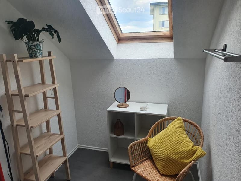 A small, cozy attic room with a skylight window, featuring a wicker chair with a yellow pillow, a wooden shelf, a small white table with a mirror, and a potted plant on the shelf.