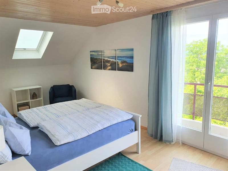 Cozy bedroom with a bed, a chair, a small shelf, and a view of the balcony. The room features a skylight and a large window with a view of greenery.