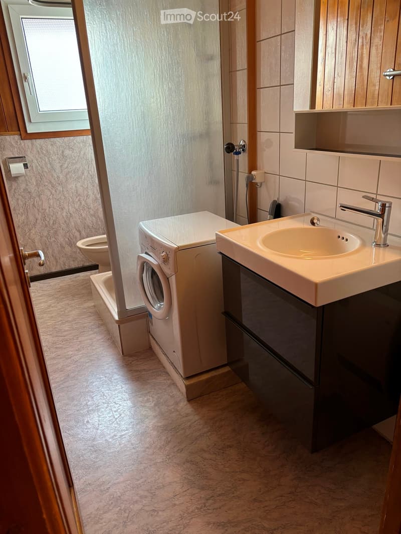 Modern bathroom with marble flooring, a white sink with a chrome faucet, a toilet, a bathtub, and a washing machine. The walls are tiled with a mix of light and dark tiles, and there is a window with wooden framing.