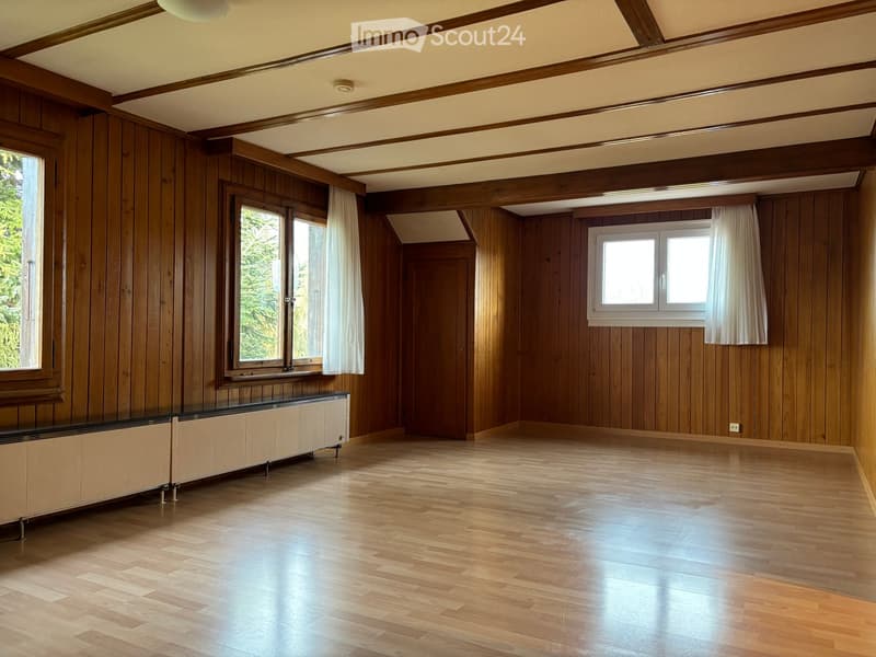 Empty room with wooden panel walls, wooden floor, large windows with white curtains, and a radiator along one wall.