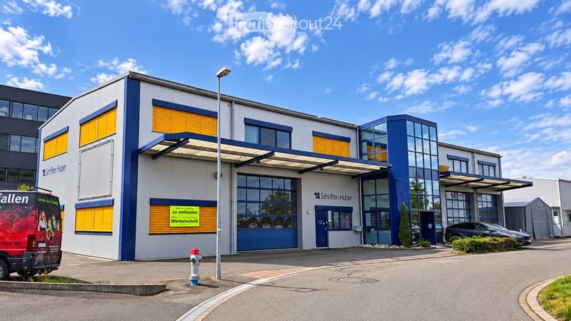 Modern industrial building with large windows, blue accents, and signage for 'Schriften Huber' and 'Werbetchnik'.