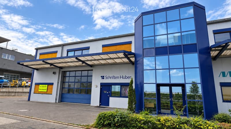 Modern commercial building with blue and white facade, large glass windows, and a covered entrance area. The building features a sign reading "Schriften Huber" and a blue garage door.