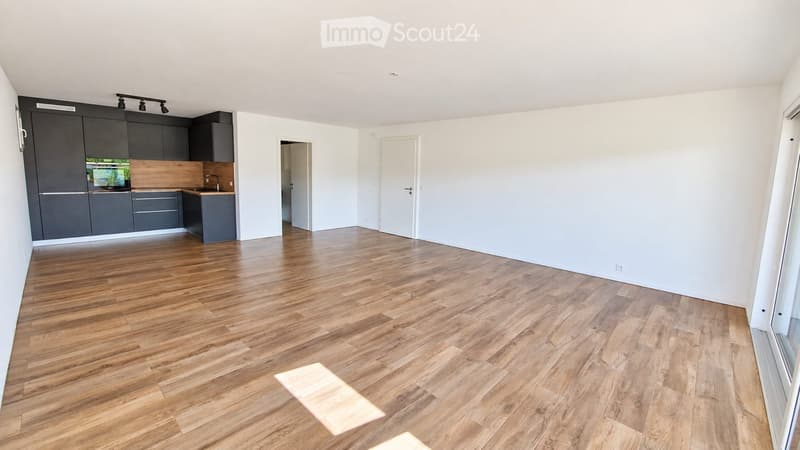 Empty room with wooden flooring, white walls, and grey kitchen cabinets visible in the background.