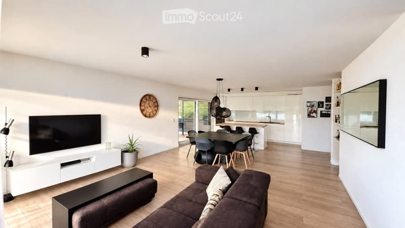 Modern living and dining area with a dark brown sofa, coffee table, flat-screen TV, dining table with chairs, and a small kitchen area with white cabinets and a countertop.