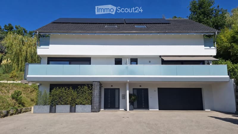 Modern two-story house with white facade, dark roof, large balcony, garage, and solar panels on the roof.