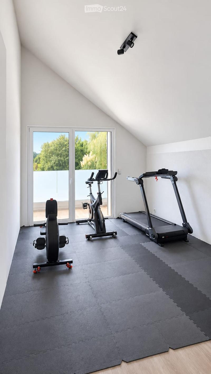 A well-lit room with a slanted ceiling, large windows, and three pieces of exercise equipment: a treadmill, an elliptical machine, and a stationary bike. The floor is covered with a black exercise mat.