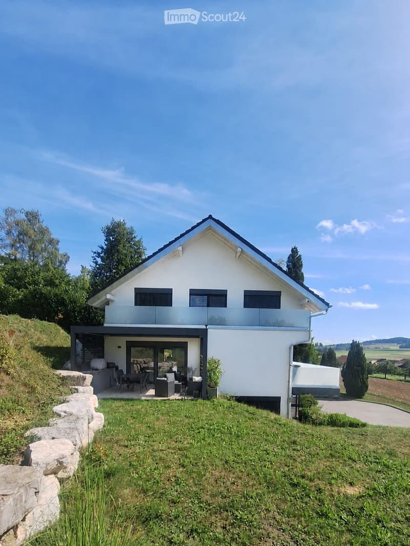 Modern two-story house with white exterior, black roof, and large windows. Features a raised ground floor, outdoor seating area, and landscaped garden.