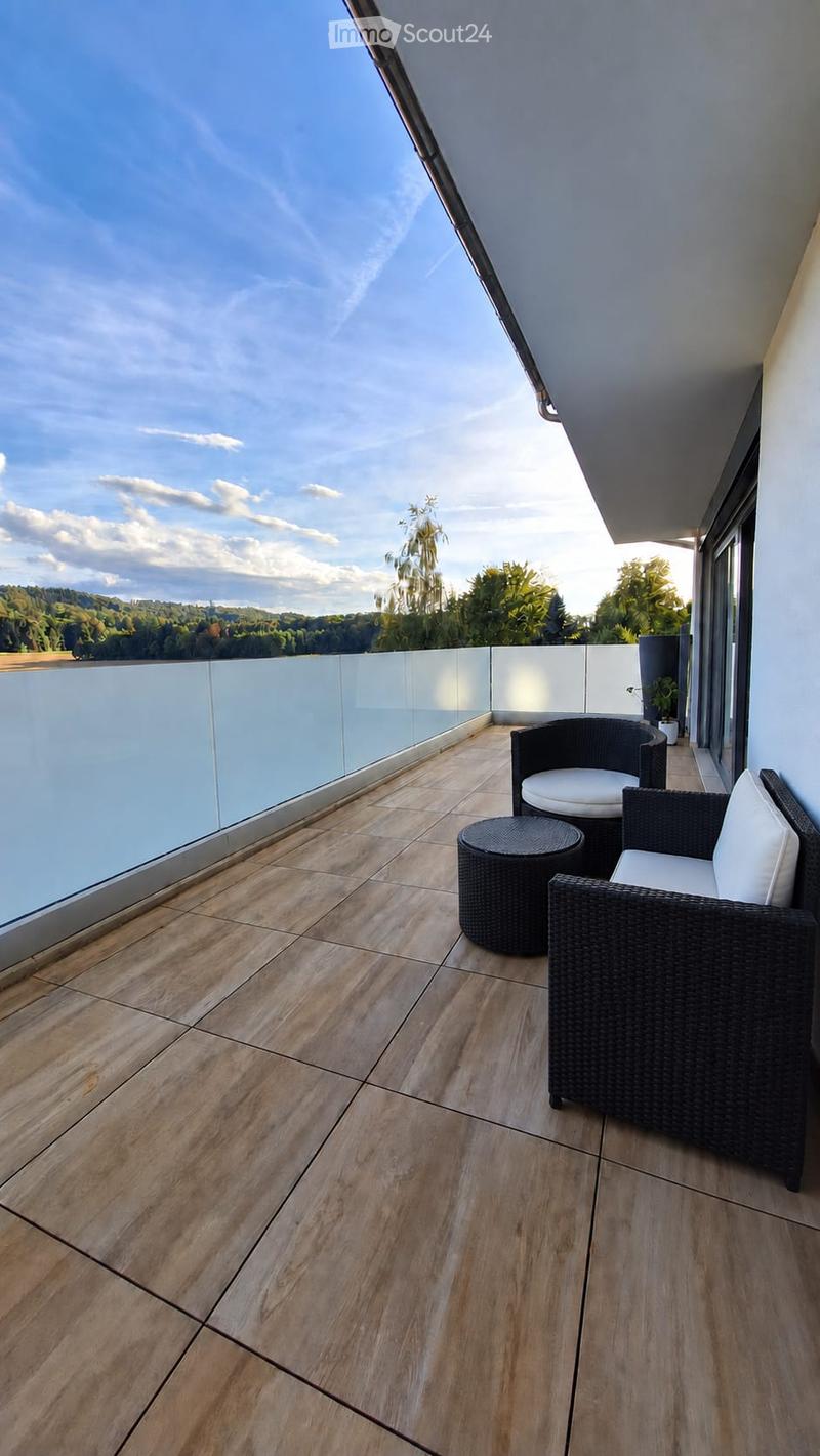 Balcony with wooden flooring, glass railing, and outdoor furniture including a wicker chair and small table.