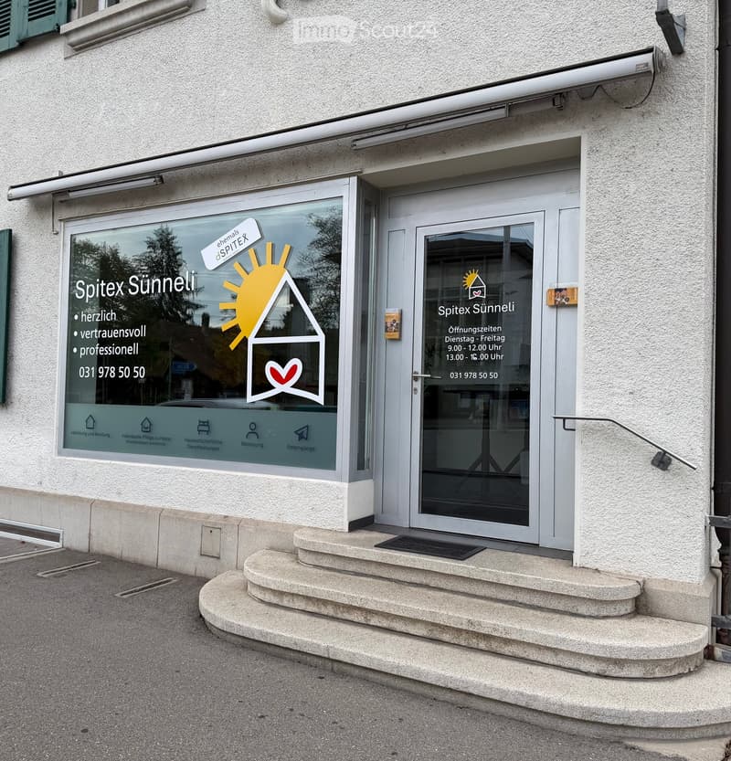 Entrance to a building with a glass door, steps, and signage for 'Spitex Sünneli' including hours and contact information.