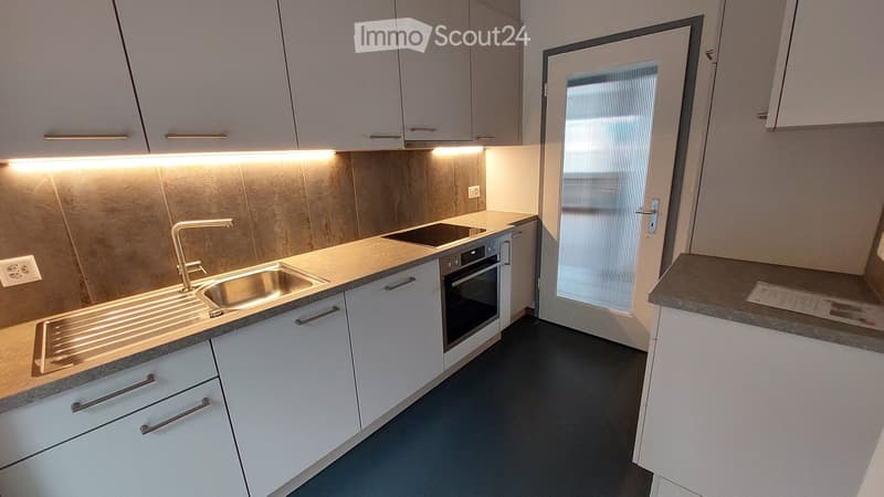 Modern kitchen with white cabinets, stainless steel sink, faucet, and built-in oven. Countertop with integrated lighting and a door leading outside.