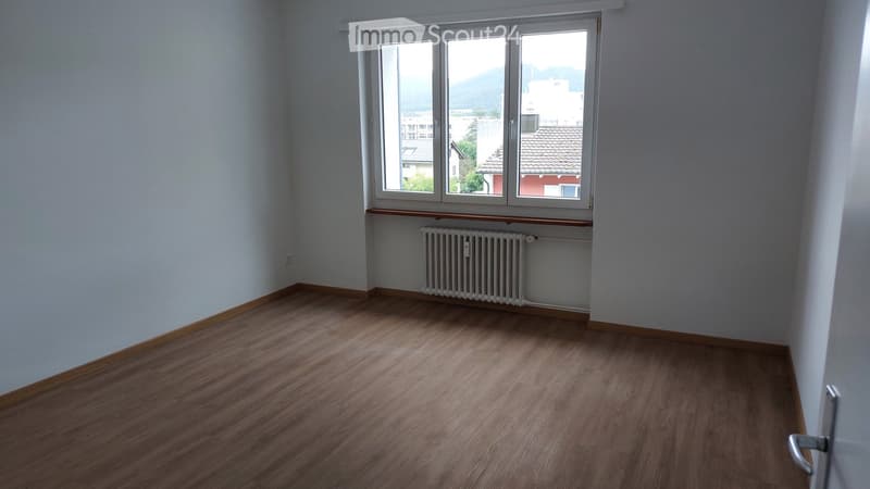 Empty room with wooden flooring, white walls, and a large window with a view of the city and mountains. A radiator is visible below the window.