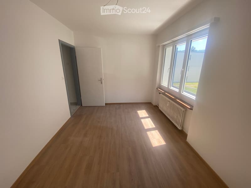 Empty room with wooden flooring, white walls, and large windows allowing natural light. A radiator is visible beneath the windows, and a door leads to an adjacent space.