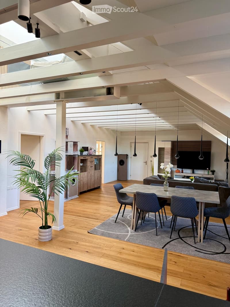 Open-concept living and dining area with wooden flooring, exposed wooden beams, pendant lights, a grey sofa, a dining table with four chairs, a cabinet, and a television on a stand.