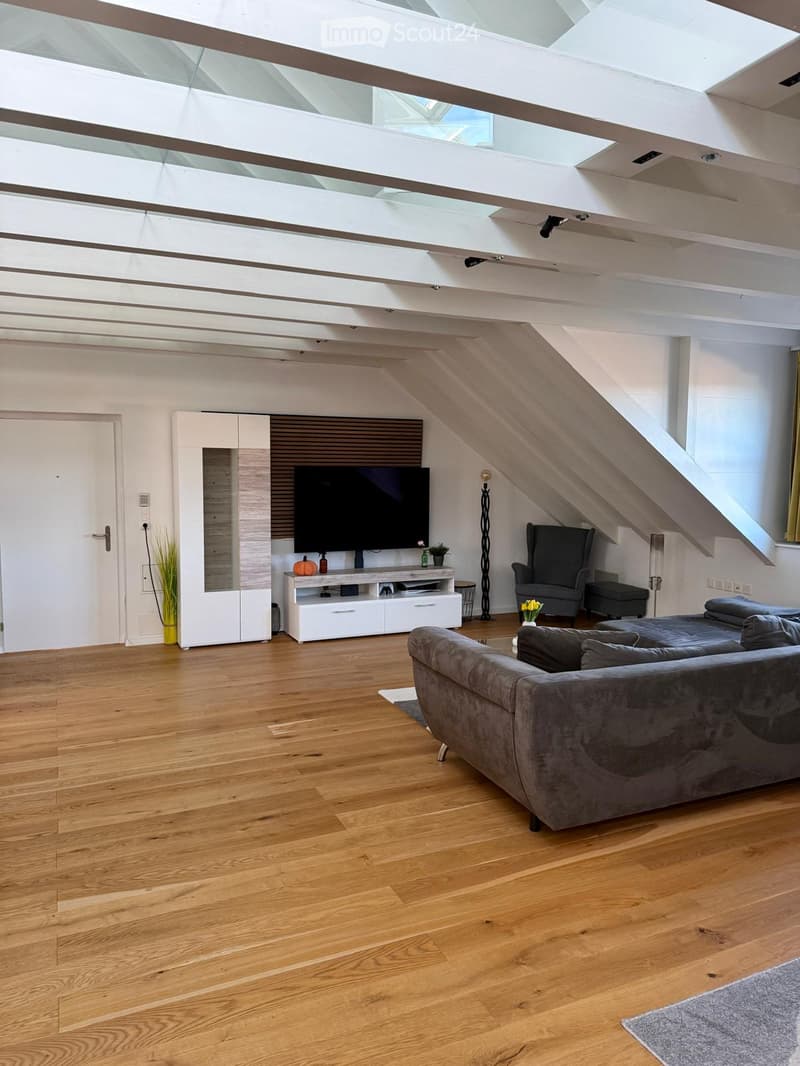 Modern living room with wooden flooring, high ceiling with skylights, grey sofa, armchair, ottoman, TV, white cabinet, and decorative items.
