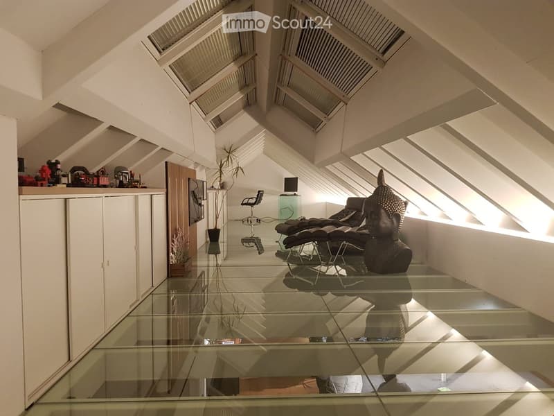 Modern, minimalist room with a glass floor, white walls, and slanted ceilings with skylights. Features a large Buddha statue, a sleek black chair, a small table, and a potted plant.