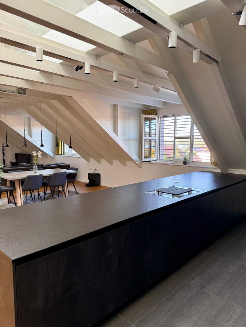 Open-plan attic space with a modern kitchen island, dining area, and living area. Features exposed wooden beams, large windows, and pendant lighting.